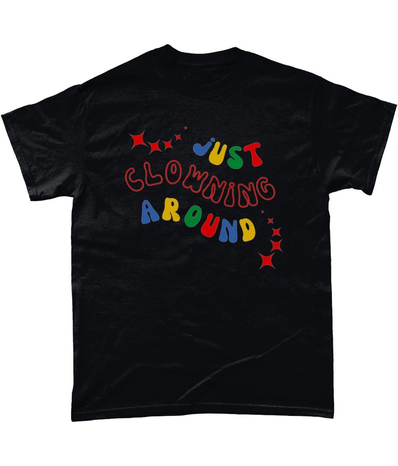 Op de afbeelding: Zwart T-shirt met de tekst "JUST CLOWNING AROUND" in kleurrijke, speelse letters. De tekst is omringd door kleine rode, gele en blauwe sterren.