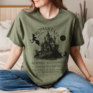 Puede incluir: Camiseta verde con una ilustraci&oacute;n en blanco y negro de un castillo embrujado, un drag&oacute;n y una bruja. El texto "ROMANTASY" est&aacute; escrito en una fuente g&oacute;tica sobre la ilustraci&oacute;n. El texto "READERS ANONYMOUS" est&aacute; escrito debajo de la ilustraci&oacute;n, junto con el texto "where love, love & magic meet".