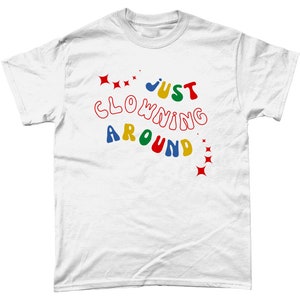 Op de afbeelding: Wit T-shirt met de tekst "JUST CLOWNING AROUND" in kleurrijke, golvende letters met rode sterren aan beide kanten.