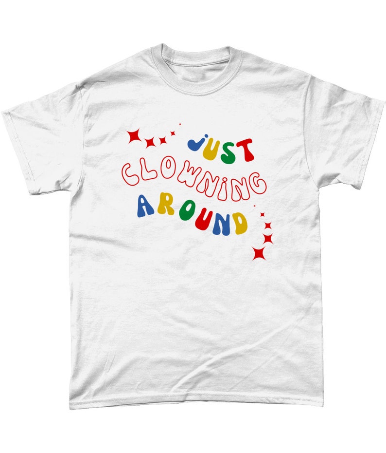 Op de afbeelding: Wit T-shirt met de tekst "JUST CLOWNING AROUND" in kleurrijke, golvende letters met rode sterren aan beide kanten.