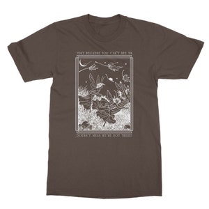 K&ouml;nnte beinhalten: Ein braunes T-Shirt mit einem wei&szlig;en Grafikdruck von Feen, die in einem Wald fliegen, mit dem Text "Just because you can't see us doesn't mean we're not there!"