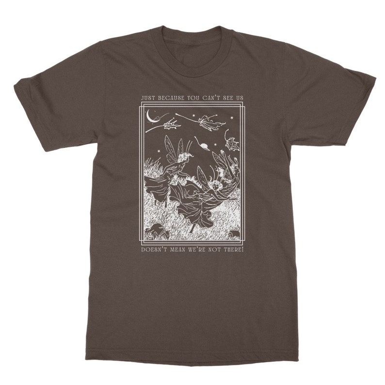 K&ouml;nnte beinhalten: Ein braunes T-Shirt mit einem wei&szlig;en Grafikdruck von Feen, die in einem Wald fliegen, mit dem Text "Just because you can't see us doesn't mean we're not there!"