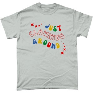 Op de afbeelding: Een lichtgrijs T-shirt met de tekst "JUST CLOWNING AROUND" in kleurrijke, golvende letters. De tekst is omringd door rode sterren.