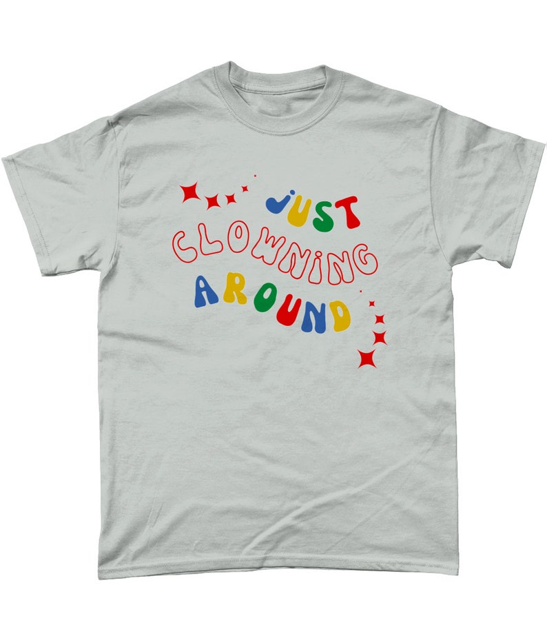 Op de afbeelding: Een lichtgrijs T-shirt met de tekst "JUST CLOWNING AROUND" in kleurrijke, golvende letters. De tekst is omringd door rode sterren.
