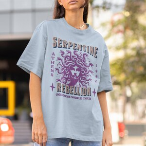 May include: A light blue t-shirt with a graphic print of a purple Medusa with snakes for hair. The text "Serpentine Rebellion" is printed below the image, with the words "Athens 1982 Goddess World Tour" printed below that.