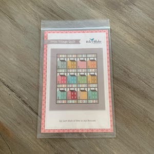 May include: A package containing a quilt pattern titled "Prim Village Quilt" by Lori Holt of Bee in my Bonnet. The pattern features a grid of colorful houses in shades of pink, blue, green, and yellow. The Riley Blake Designs logo is visible.