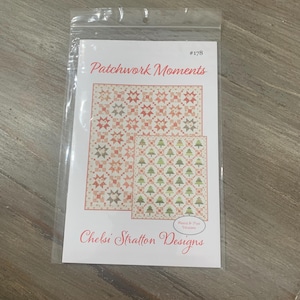 May include: A clear plastic bag containing a pattern for a quilt. The pattern is titled "Patchwork Moments" and features two quilt designs with star and Christmas tree patterns in shades of peach, green, and white. The designer's name, Chelsi Stratton Designs, is also visible.