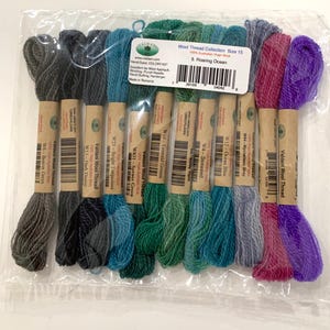 May include: A collection of twelve skeins of Valdani wool thread in the Roaring Ocean color palette, packaged in a clear plastic bag. The threads range in color from gray and dark blue to various shades of green, teal, pink, and purple.