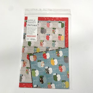 May include: A sewing pattern for a quilt featuring a red, white, and blue apple design. The pattern is called "Apple Dandy" and the pattern number is "PAT068". The pattern is by BasicGrey Sewing Patterns.
