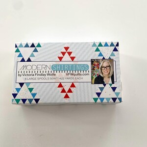 May include: A box of Modern Shirtings fabric by Victoria Findlay Wolfe. The box features a white background with a pattern of blue and green triangles. The box also has a red and white triangle pattern. The box has a label that says "Modern Shirtings by Victoria Findlay Wolfe VFQuilts.com 4 Large Spools 50WT 1422 Yards Each".