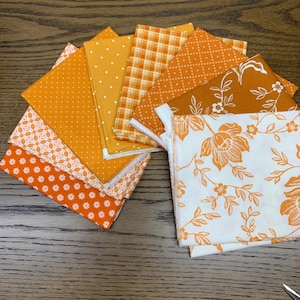 Peut inclure: Une collection d'échantillons de tissu carrés disposés en éventail, présentant divers motifs orange et blancs, notamment des pois, des carreaux, des petites fleurs, des formes géométriques et un grand imprimé floral sur fond blanc.