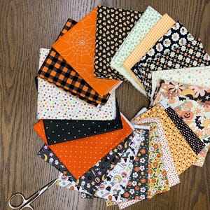 May include: A collection of folded fabric squares arranged in a circular fan pattern, featuring various Halloween-themed prints including orange spiderwebs, black and orange buffalo plaid, ghosts, pumpkins, bats, skulls, and floral patterns.