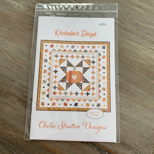 May include: A quilt pattern titled "October Days" by Chelsi Stratton Designs. The pattern features a central pumpkin design surrounded by autumn leaves and cross motifs in shades of orange, brown, and cream. The pattern is packaged in a clear plastic sleeve.