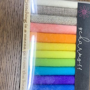 May include: A clear plastic package containing twelve rolled fabric strips in a rainbow gradient of colors including white, gray, pink, orange, yellow, lime, green, teal, purple, and blue. The packaging features the text #charms11 and Stitching is a message from the heart.