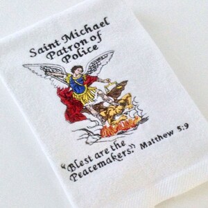 May include: White hand towel featuring an embroidered image of Saint Michael, the patron saint of police, with a sword and wings. Text reads "Saint Michael Patron of Police" and "Blest are the Peacemakers!" Matthew 5:9.