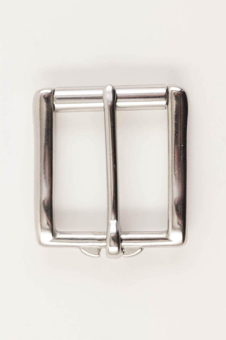 May include: A shiny, silver-colored belt buckle with a rectangular frame and a central bar. The buckle has a classic design, suitable for various belts. The metal has a polished finish, reflecting light.