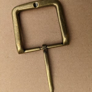 Op de afbeelding: Een messingkleurige metalen gesp met een rechthoekig frame en een enkele pen.