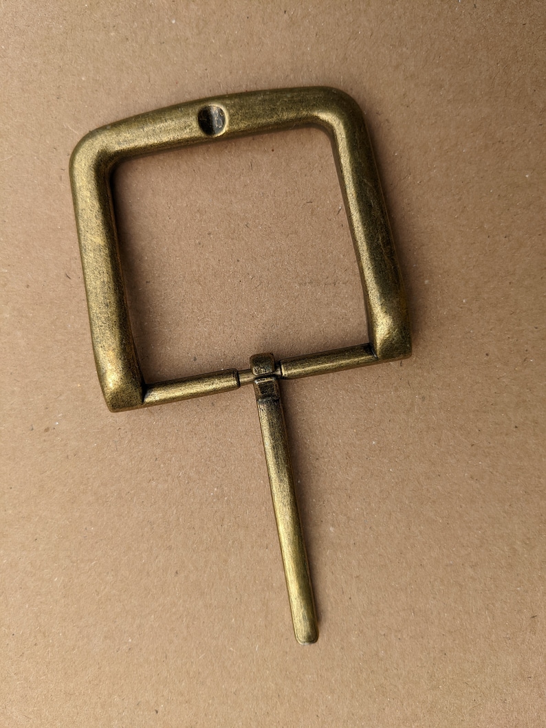 Op de afbeelding: Een messingkleurige metalen gesp met een rechthoekig frame en een enkele pen.