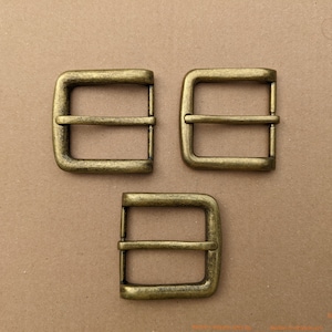 Op de afbeelding: Drie antieke goudkleurige metalen riemgespen. Elke gesp heeft een rechthoekige vorm met een enkele, gecentreerde, horizontale balk.
