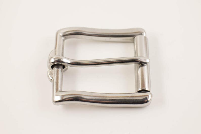 May include: A shiny, silver-colored metal belt buckle. The buckle has a rectangular shape with a curved top and bottom bar. The design includes a central bar and a pin for securing a belt. The buckle is shown against a white background.