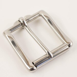 May include: A shiny, silver-colored belt buckle with a rectangular frame and a single, centered bar. The buckle is made of metal and has a polished finish, reflecting light. The design is simple and classic.