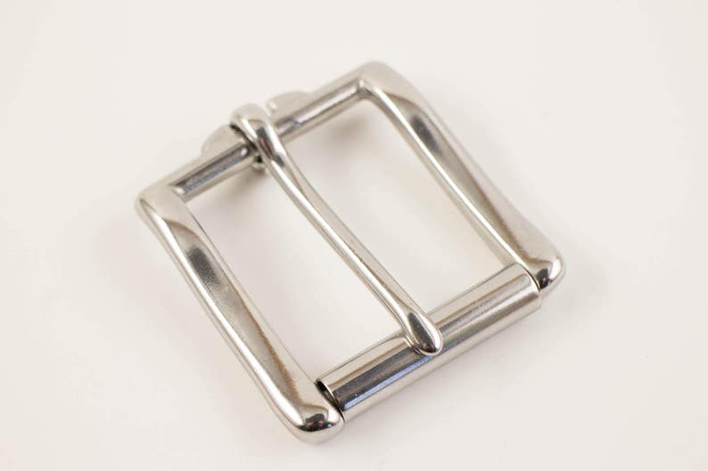 May include: A shiny, silver-colored belt buckle with a rectangular frame and a single, centered bar. The buckle is made of metal and has a polished finish, reflecting light. The design is simple and classic.