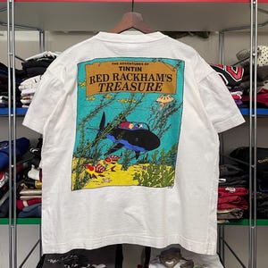 May include: White t-shirt featuring a colorful graphic from "The Adventures of Tintin: Red Rackham's Treasure." The design depicts an underwater scene with a shark, fish, and coral. The text "Red Rackham's Treasure" is displayed in a brown banner.
