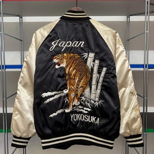 Può includere: Giacca bomber in raso nero con maniche color crema. La schiena presenta una tigre ricamata, bambù e le parole "Japan" e "Yokosuka" in scrittura bianca. La giacca ha un bordo a righe bianche e nere.