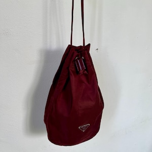Puede incluir: Un bolso burdeos con cordón y correa larga, colgado contra una pared blanca. El bolso presenta un pequeño logotipo plateado y un detalle de cuero. El bolso está hecho de un material liso, posiblemente nailon.