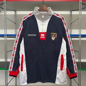 May include: A navy blue and white long-sleeve athletic top with red accents. The top features a white collar with a zipper, the word "KENWOOD", and the "errea" logo. The sleeves have a repeating pattern of the word "errea".