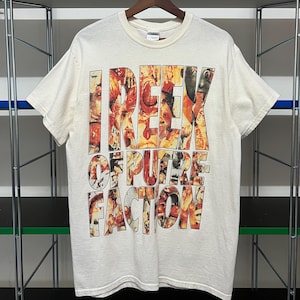 Puede incluir: Camiseta color crema con el texto "I REEK OF PURE FACTION" en una fuente audaz y multicolor de estilo collage. La camiseta está colgada en una percha de madera sobre un fondo neutro.