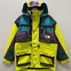 Puede incluir: Una chaqueta North Face amarilla, verde y morada con múltiples bolsillos y capucha. La chaqueta tiene el logotipo de North Face en el pecho izquierdo.