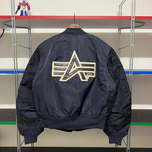 以下が含まれることがあります： ネイビーブルーのボンバージャケット、黒い袖付き。背面には「USA」の文字が入った、大きく金と黒のロゴがあります。ジャケットは木製のハンガーに吊るされています。