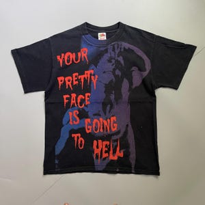 Peut inclure: Un t-shirt noir à manches courtes avec un motif représentant une personne la main sur la tête dans des tons violets et bleus. Le texte "YOUR PRETTY FACE IS GOING TO HELL" est imprimé en rouge avec un effet dégoulinant sur le devant.