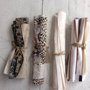 Könnte beinhalten: Vier Stoffrollen, mit Bindfaden gebunden, liegen vor einer weißen, verwitterten Holzoberfläche. Die Stoffe zeigen verschiedene Muster, darunter Animal-Prints und Streifen, in neutralen Tönen von Beige, Schwarz und Weiß.