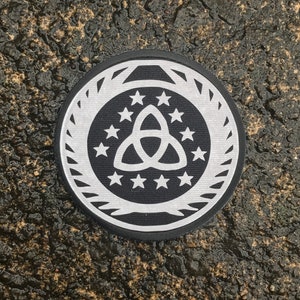 May include: A circular black and white patch featuring a stylized triquetra symbol surrounded by stars and a decorative border. The patch is set against a dark, textured background.