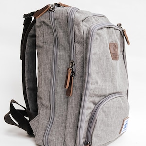 May include: A heather gray backpack featuring multiple zippered compartments, brown leather-like zipper pulls, and black adjustable shoulder straps. A brown rectangular patch with a logo is on the front, along with a small white rectangular label.