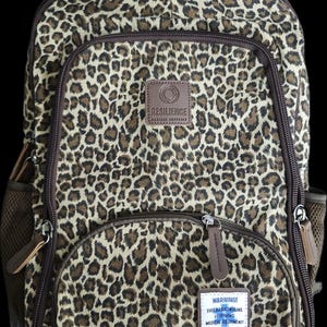 Peut inclure: Un sac à dos à motif léopard avec des bordures marron, doté d'un compartiment supérieur zippé et d'une poche avant. Un écusson marron indique RESILIENCE MEDICAL SUPPLIES. Une étiquette blanche en bas indique WARNING! THIS BAG CONTAINS LIFESAVING MEDICAL EQUIPMENT.