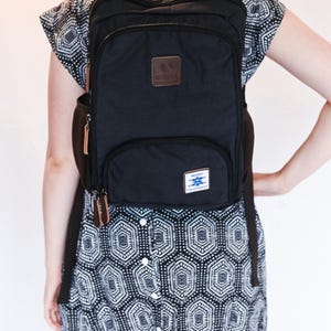 May include: A black backpack with multiple zippered compartments, brown leather-like zipper pulls, and a brown rectangular patch on the upper front. A small white rectangular label with blue text is attached to the lower front pocket.