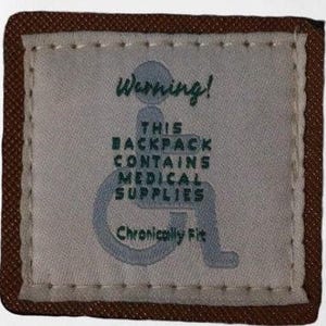 Pode incluir: Um remendo de tecido castanho com costuras brancas ao redor das bordas. O remendo tem um fundo branco com texto verde que diz "Aviso! Esta mochila contém suprimentos médicos Chronically Fit". Um símbolo de cadeira de rodas estilizado está no centro do remendo.