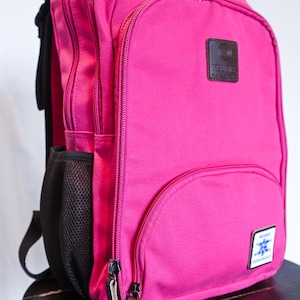 May include: A bright pink backpack featuring multiple zippered compartments, a black mesh side pocket, and two rectangular patches on the front. One patch is dark brown with text, and the other is white with a blue graphic and text.