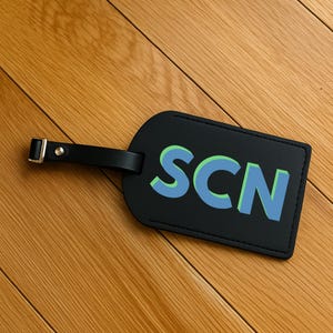 May include: Black luggage tag with the letters "SCN" in blue and green. The tag has a black leather strap with a silver buckle. The tag is rectangular with rounded corners and is lying on a wooden surface.