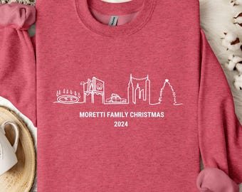 May include: A red sweatshirt with a white outline of a city skyline and a Christmas tree. The text "Moretti Family Christmas 2024" is printed below the skyline.