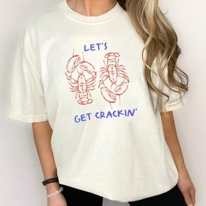 May include: Off-white t-shirt featuring a red lobster design and the blue text "LET'S GET CRACKIN'". The shirt has a relaxed fit and short sleeves, suitable for everyday wear.
