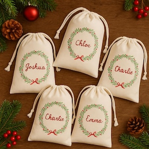 Puede incluir: Cinco bolsas de regalo crema con cordón, cada una con un nombre en escritura roja. Cuentan con una corona verde con bayas rojas y un lazo rojo. Tema navideño.