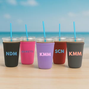 May include: Five iced coffee cups with blue straws and personalised sleeves. The sleeves are black, pink, purple, red, and grey, each with a different three-letter monogram. The background is a beach scene with blue water and sky.