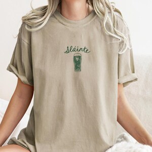 May include: A light beige t-shirt with a green graphic of a glass of beer and the text "Sl&aacute;inte" printed on the front.