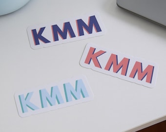 May include: Three rectangular stickers with the letters "KMM" in different color schemes. One sticker has blue letters with a pink outline, another has pink letters with a blue outline, and the last has light blue letters with a green outline.