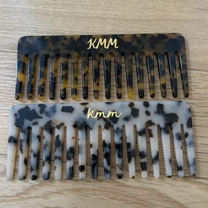 May include: Two rectangular hair combs. One is a tortoiseshell pattern with the gold letters "KMM". The other comb is white with black spots and the gold letters "kmm". Both combs have evenly spaced teeth.