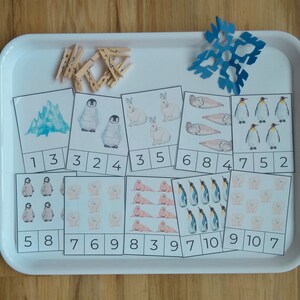 Può includere: Un vassoio bianco con un ritaglio a forma di fiocco di neve e 10 carte con diversi numeri di animali, tra cui pinguini, foche e orsi polari. I numeri da 1 a 10 sono scritti sulle carte. Ci sono anche delle mollette di legno sul vassoio.